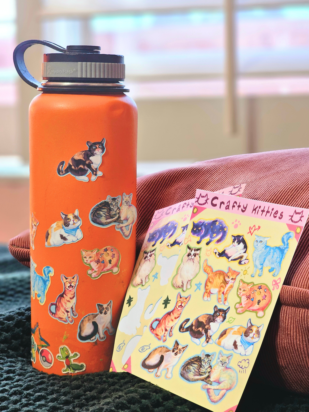 Orange water bottle with cat stickers next to a sheet of cat-themed stickers on a couch.