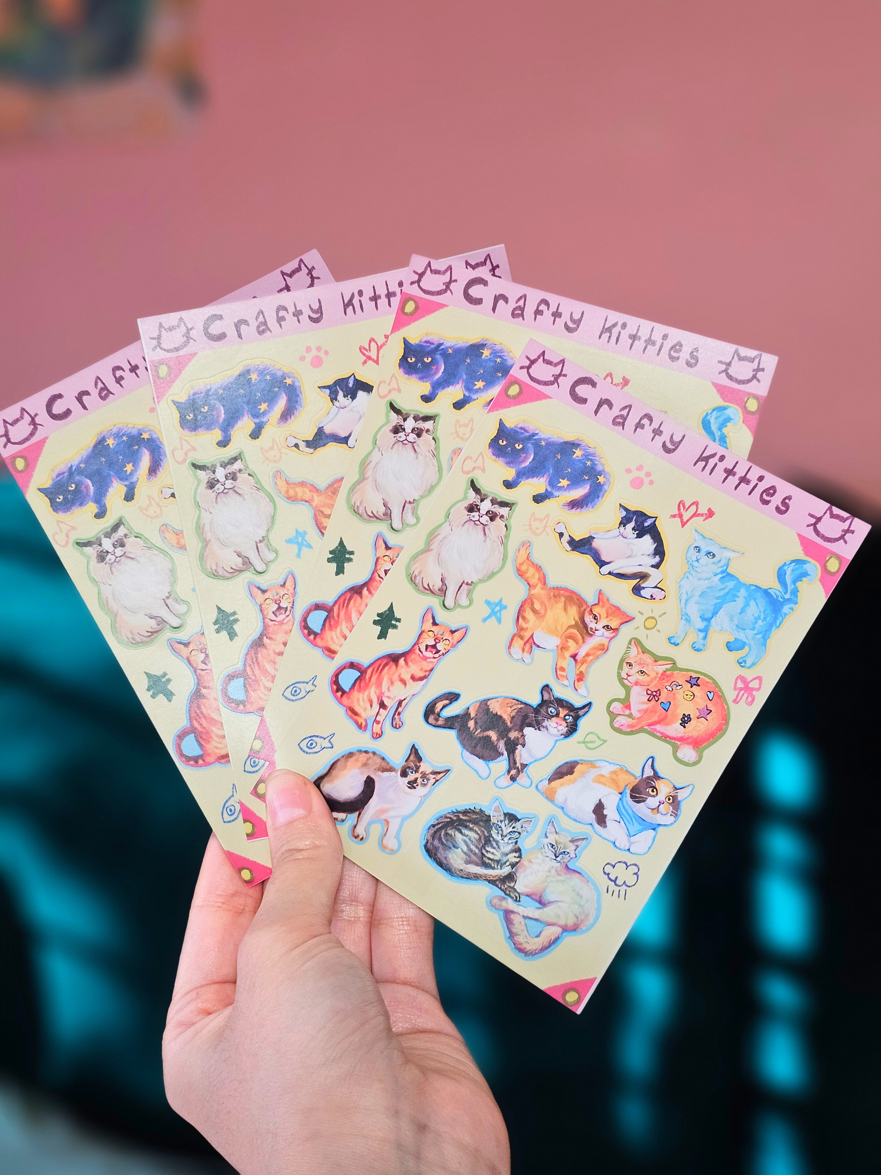 Hand holding sheets of 'Crafty Kitties' sticker sheets with colorful cat illustrations on a pink background.