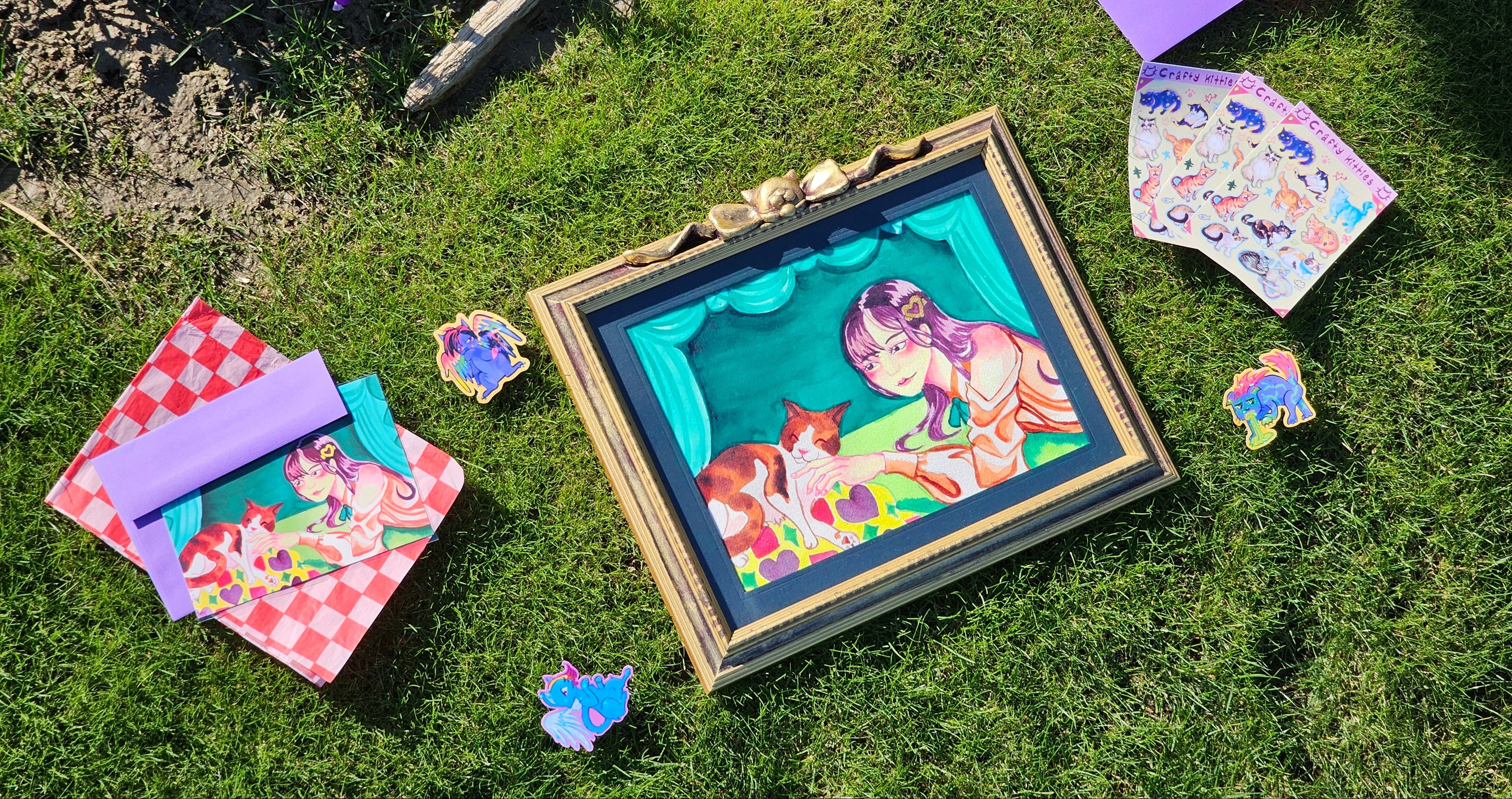 Colorful art pieces on grass, including stickers, postcards, and a painting.