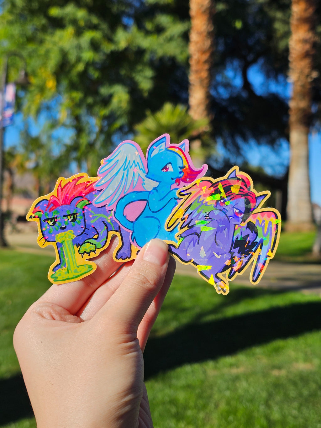 Three colorful die-cut cat stickers held by a hand with a park background, depicting the stickers (each sold separately) in the Cringe Cats sticker collection.