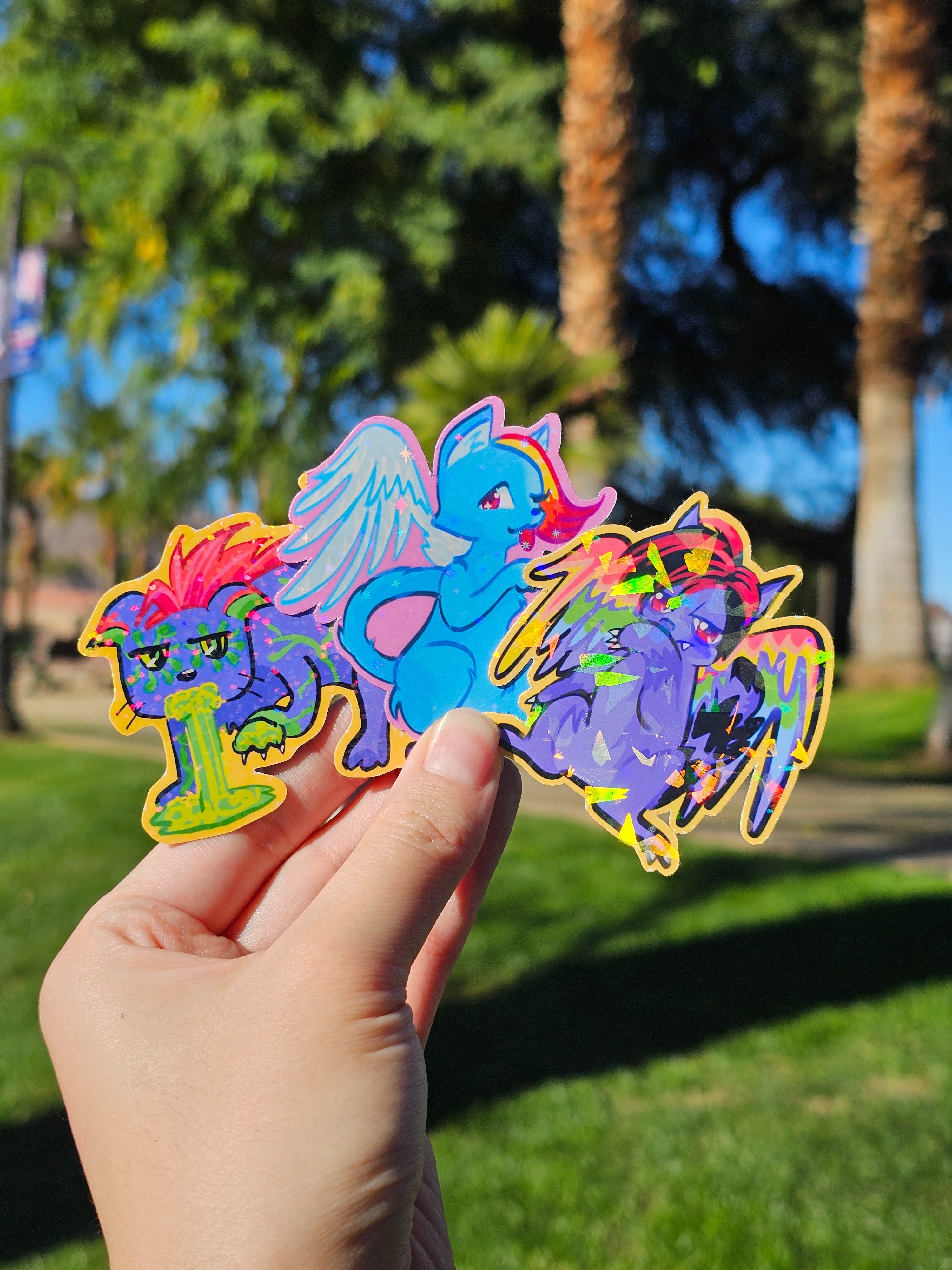 Three colorful die-cut cat stickers held by a hand with a park background, depicting the stickers (each sold separately) in the Cringe Cats sticker collection.