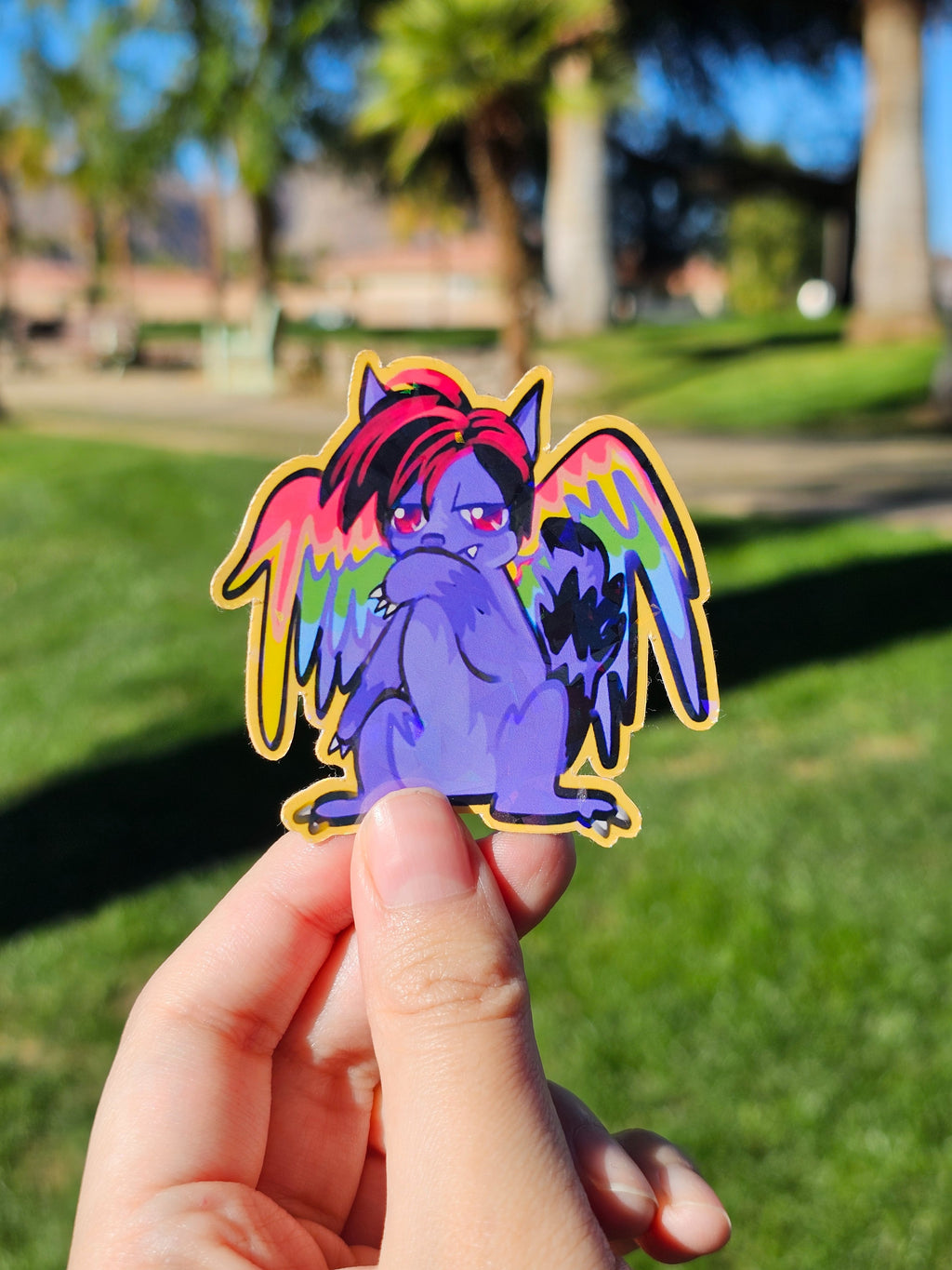 Hand holding a colorful die-cut sticker of a rainbow winged cat with red and black scene hair and captivating red eyes in a blurred park background