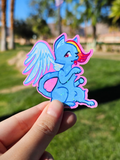Hand holding a die-cut sticker of a blue winged cat with multi-colored hair in a park in the background